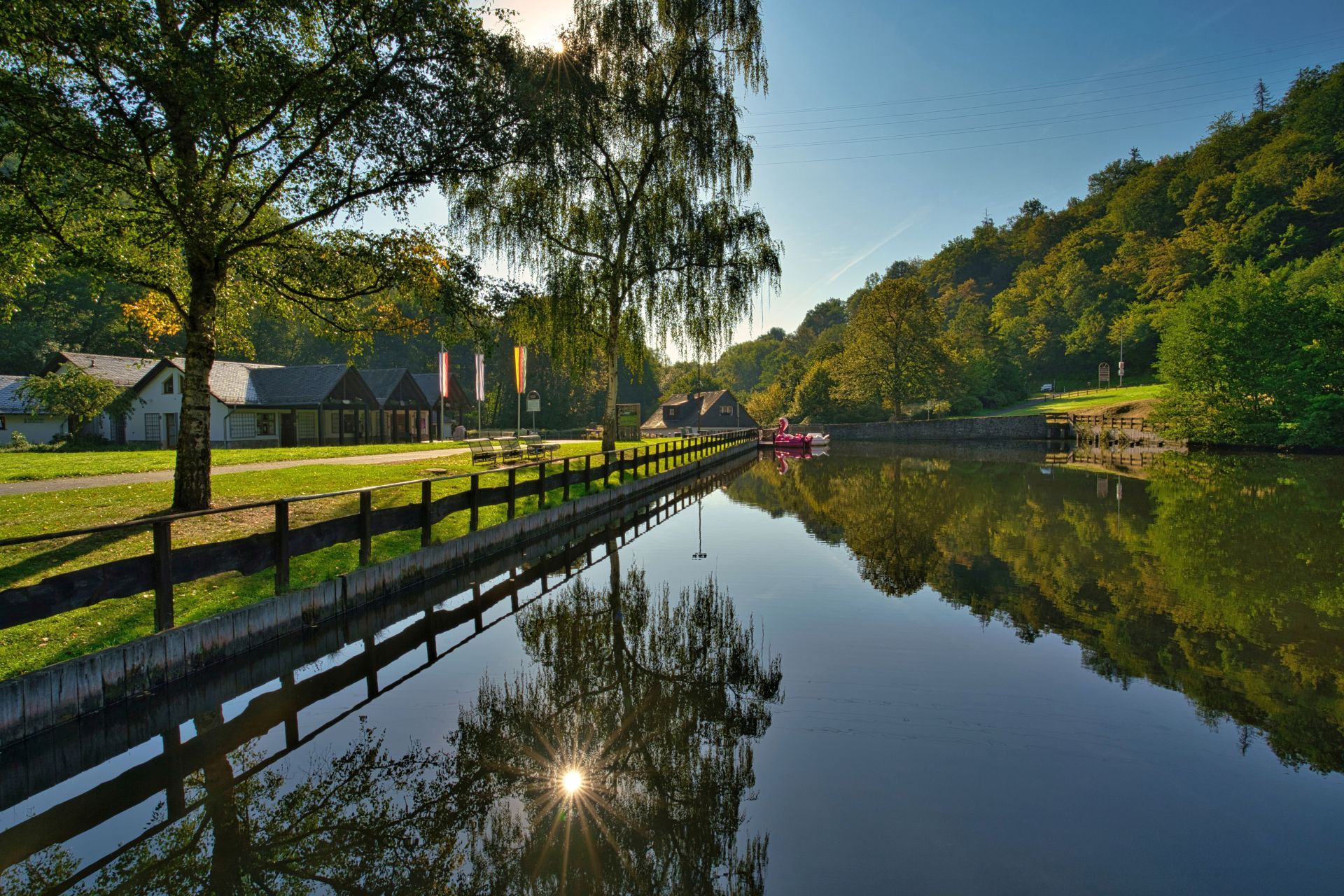 The photo shows a view over the Kallwiesweiher pond. The buildings of the visitor center can be seen in the background on the left and the Weiherschleife building next to it.