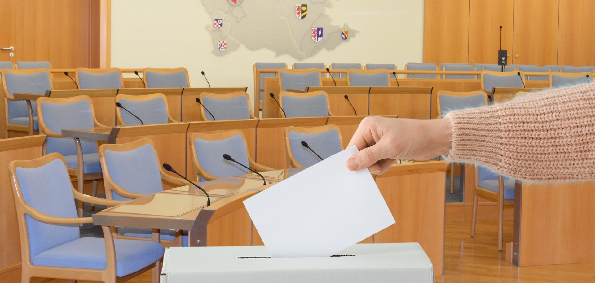 The photo shows an arm inserting a ballot paper into a ballot box. The meeting room of the Idar-Oberstein city council can be seen in the background.