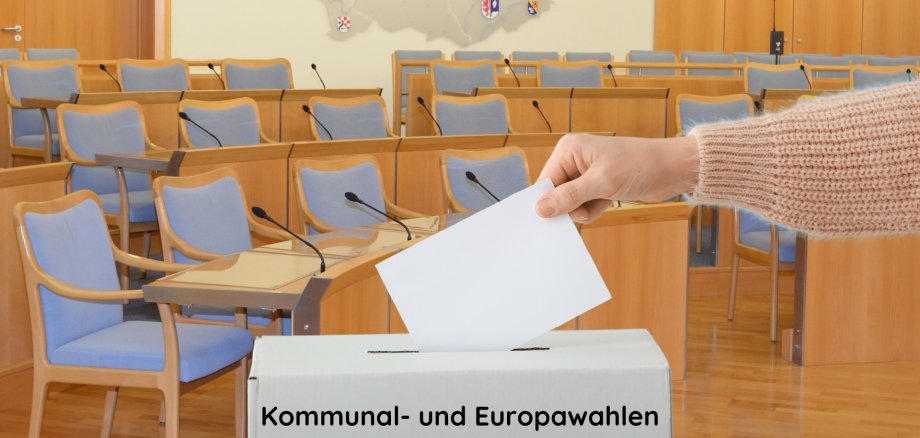 The photo shows an arm inserting a ballot paper into a ballot box. The meeting room of the Idar-Oberstein city council can be seen in the background.