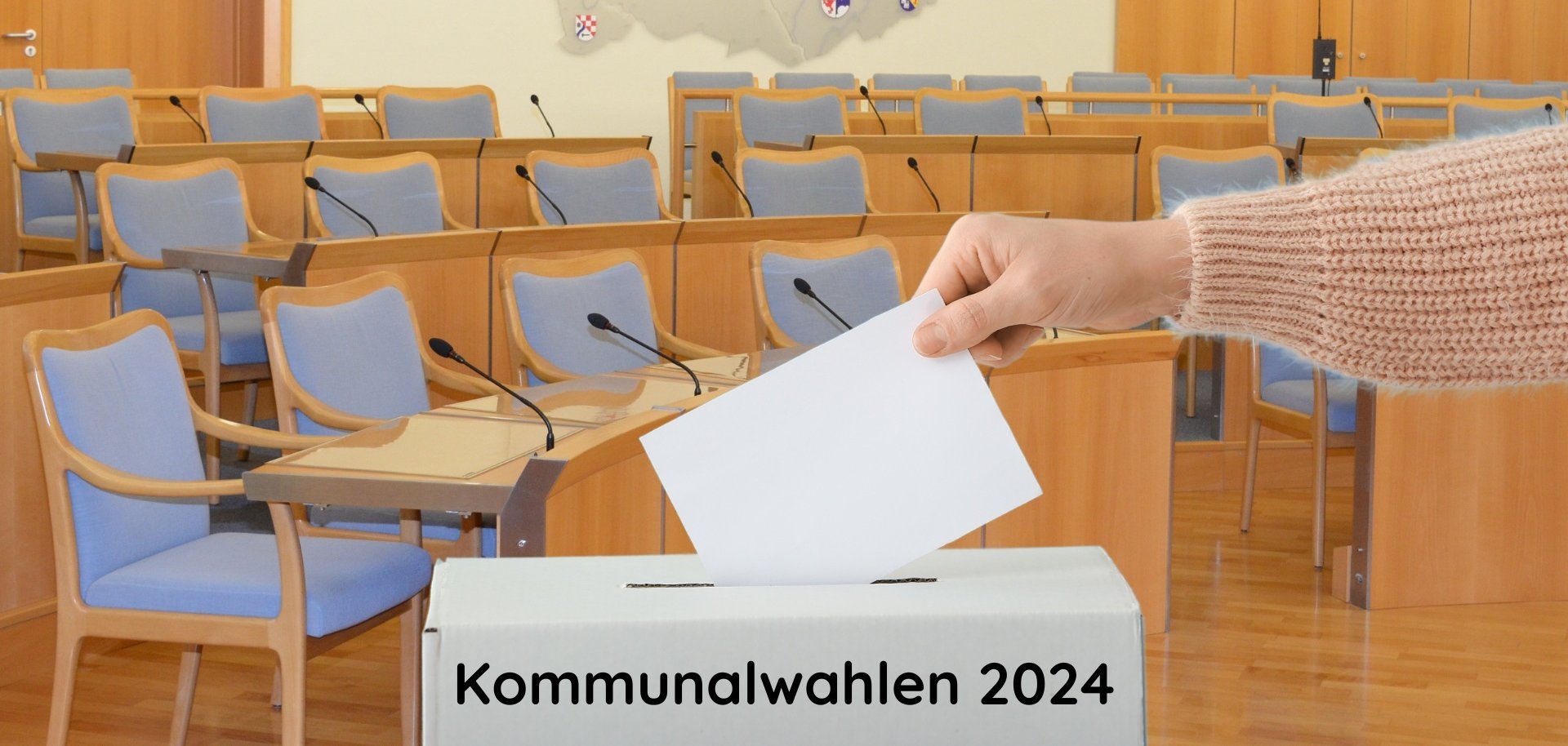 The photo shows an arm inserting a ballot paper into a ballot box. The meeting room of the Idar-Oberstein city council can be seen in the background.