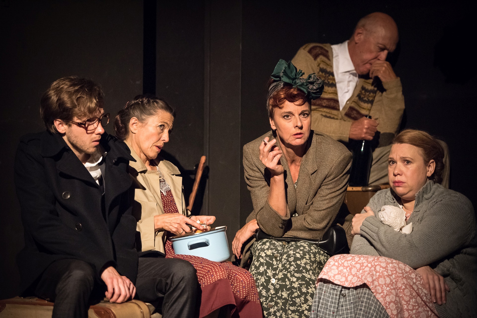 The scene photo shows five actors and actresses sitting huddled together in a cellar.