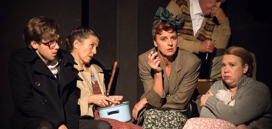 The scene photo shows five actors and actresses sitting huddled together in a cellar.