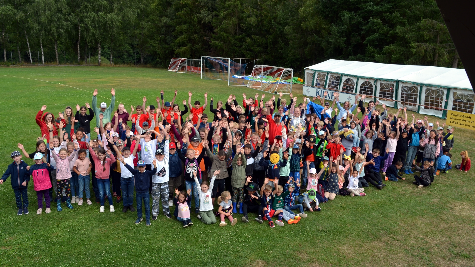 The photo shows the children of the city recreation with supervisors and visitors. They are all standing on the sports field and waving at the camera.