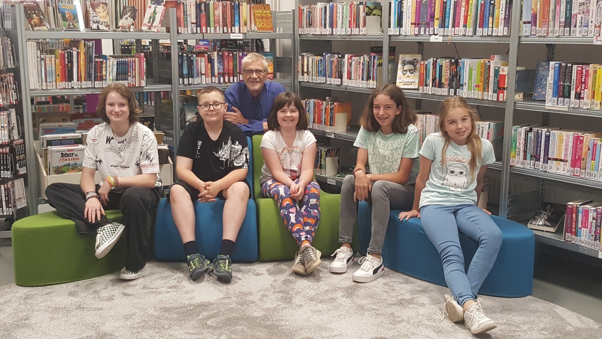 The photo shows the participants of the writing workshop sitting next to each other on colorful armchairs with Jörg Staiber behind them.