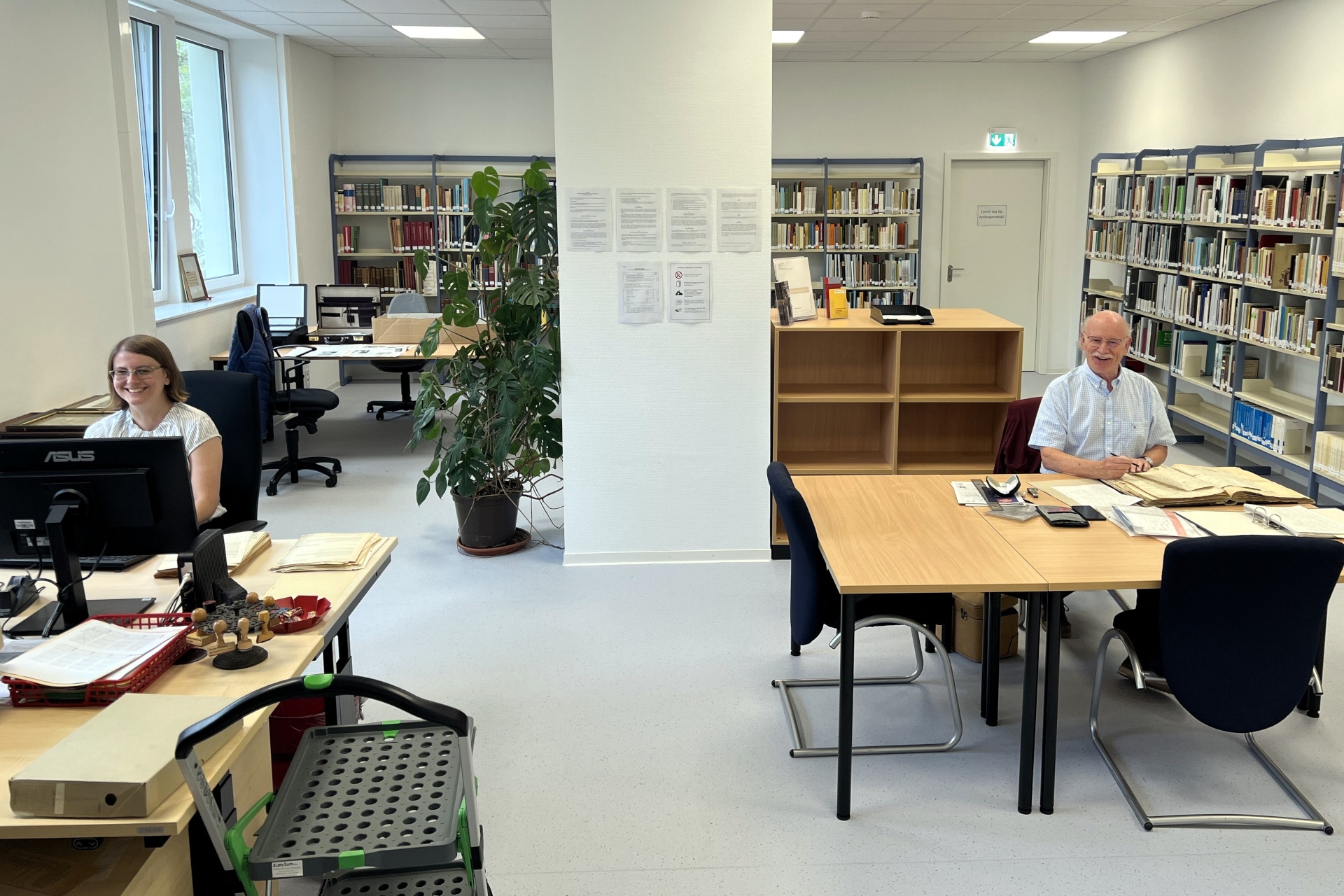 The photo shows a view of the new reading room with shelves and tables. Dr. Svenja Müller is sitting at a desk on the left-hand side of the picture and Dieter Jerusalem is sitting at a table with old documents on the right-hand side.