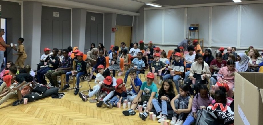 The photo shows many children sitting in a semicircle on the floor and watching the movie.