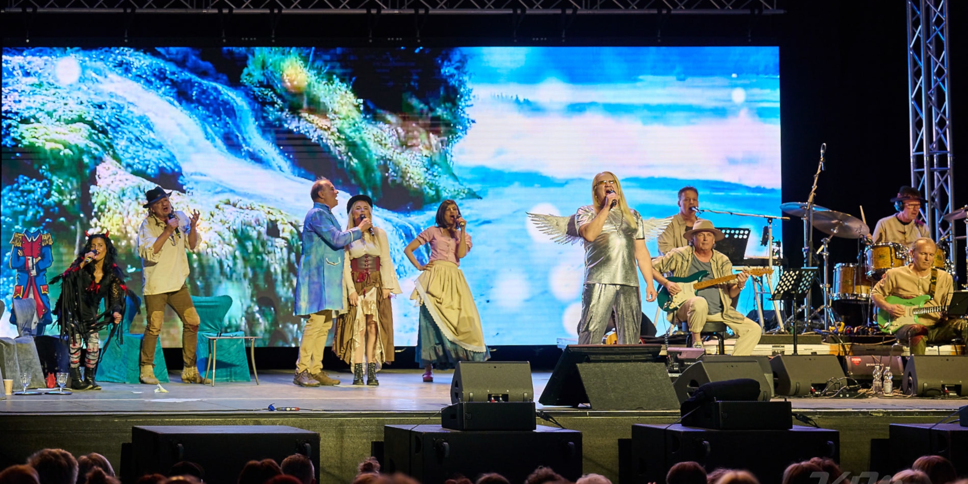 The photo shows the six performers in the left and center of the picture. They are standing next to each other on stage and singing. On the right is the band Die Crackers, who are playing live.