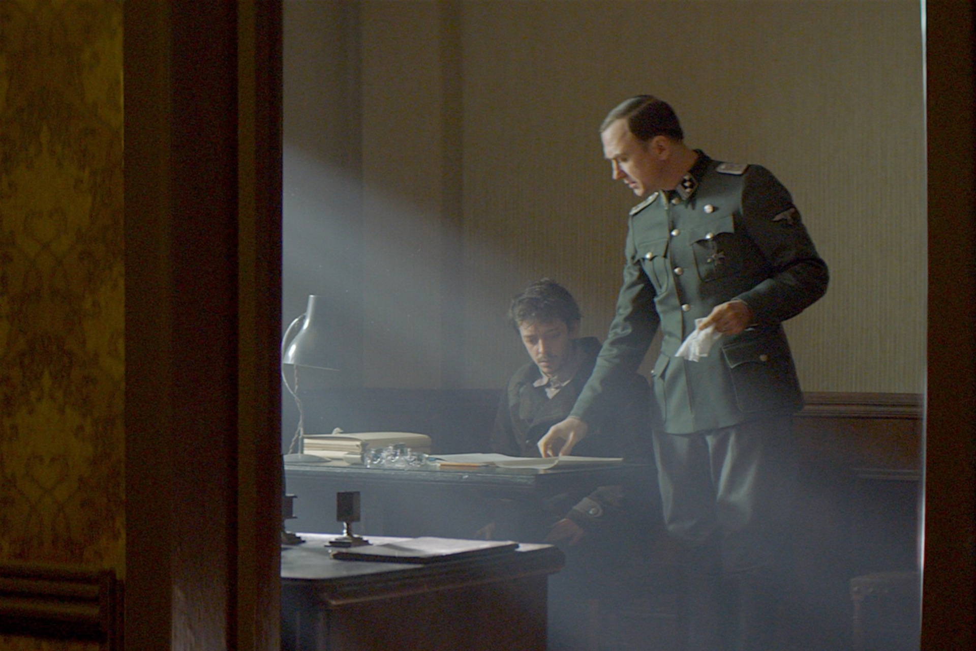 The photo shows two men in a dark room. One is sitting at a desk. The other is standing next to him, leafing through a book lying on the desk.