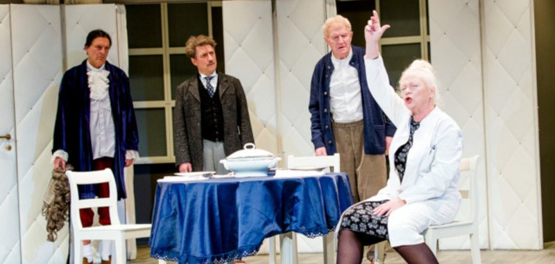 The scene photo from the play shows an actress sitting at a table and gesticulating wildly. Three actors stand behind the table and look at her, puzzled.