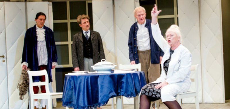 The scene photo from the play shows an actress sitting at a table and gesticulating wildly. Three actors stand behind the table and look at her, puzzled.