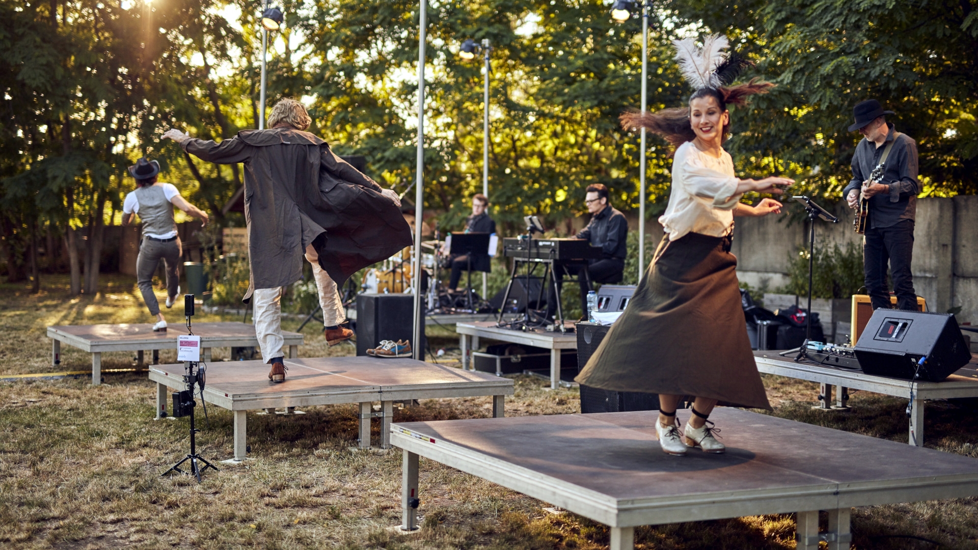 The photo shows three dancers from the company dancing individually on stage platforms outdoors. Behind them are musicians who are also placed individually on stage platforms.