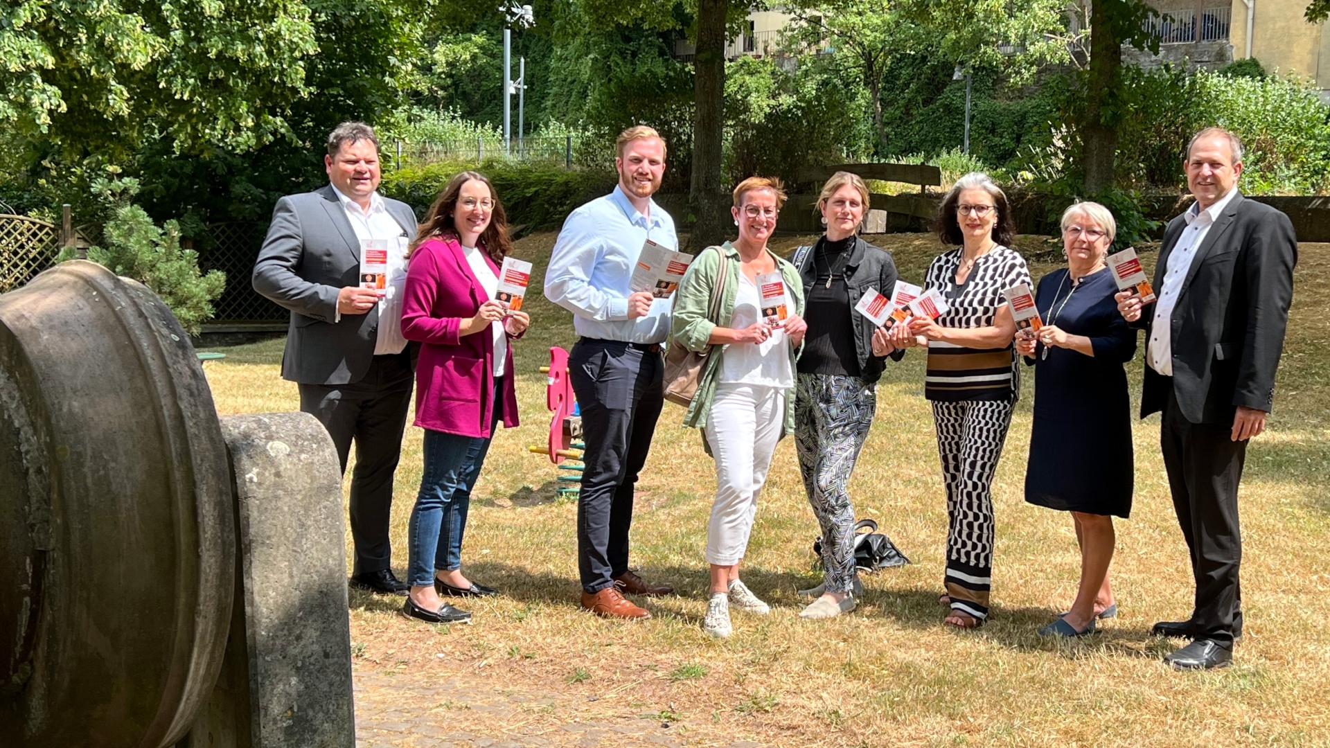 The photo shows eight people standing in a green area next to an old grindstone and holding event flyers.