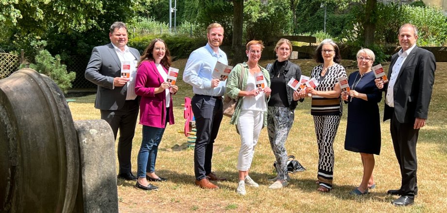 The photo shows eight people standing in a green area next to an old grindstone and holding event flyers.