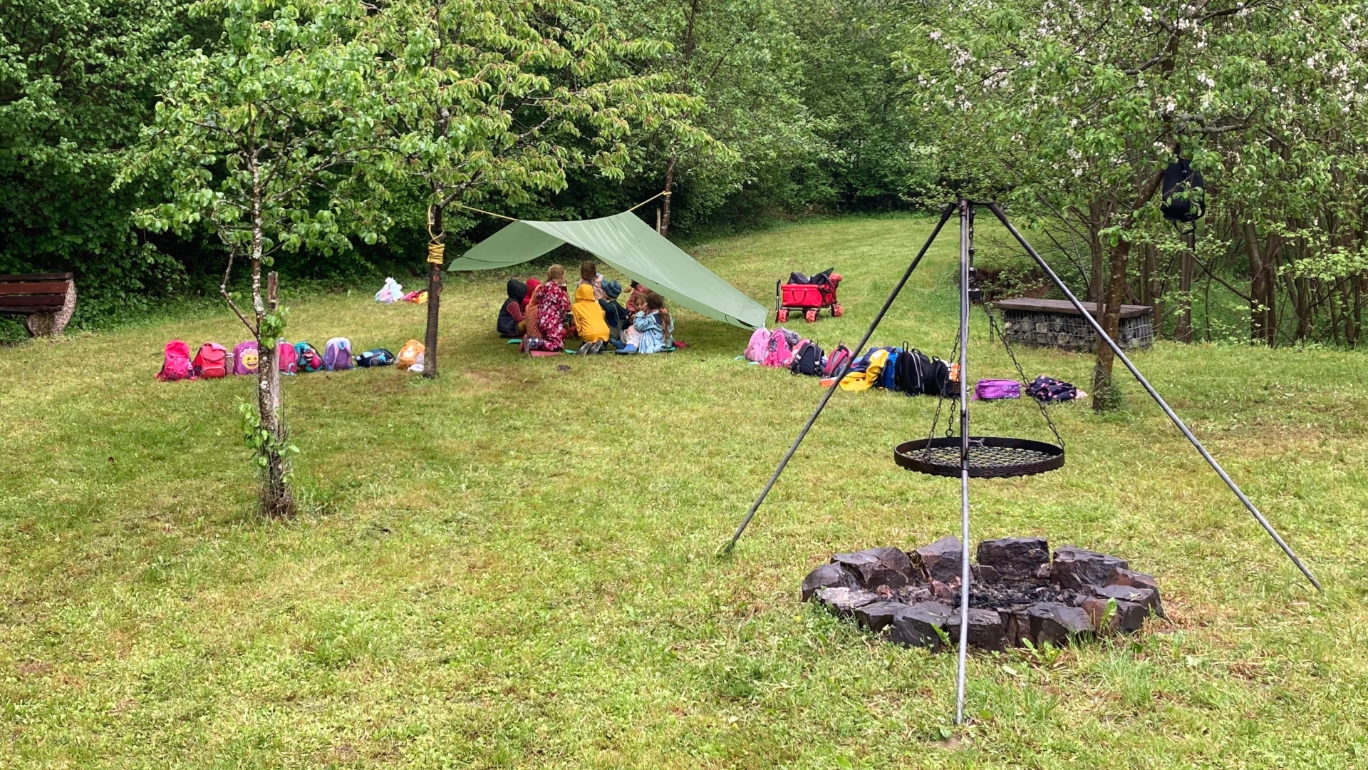The photo shows the children in the background, sitting under a tarpaulin on a meadow. A tripod with a swivel grate can be seen in the foreground.