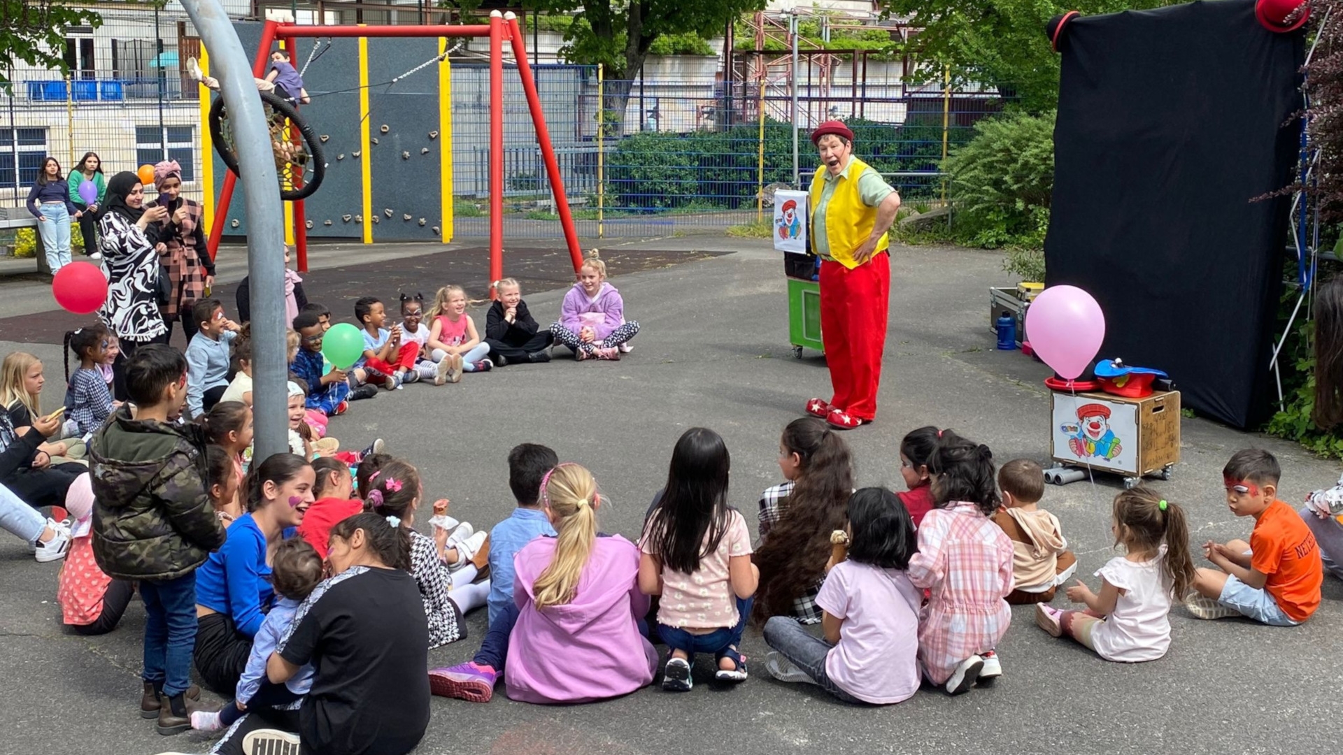 The photo shows a large group of children sitting in a semi-circle in front of the clown Kuni on the event site.
