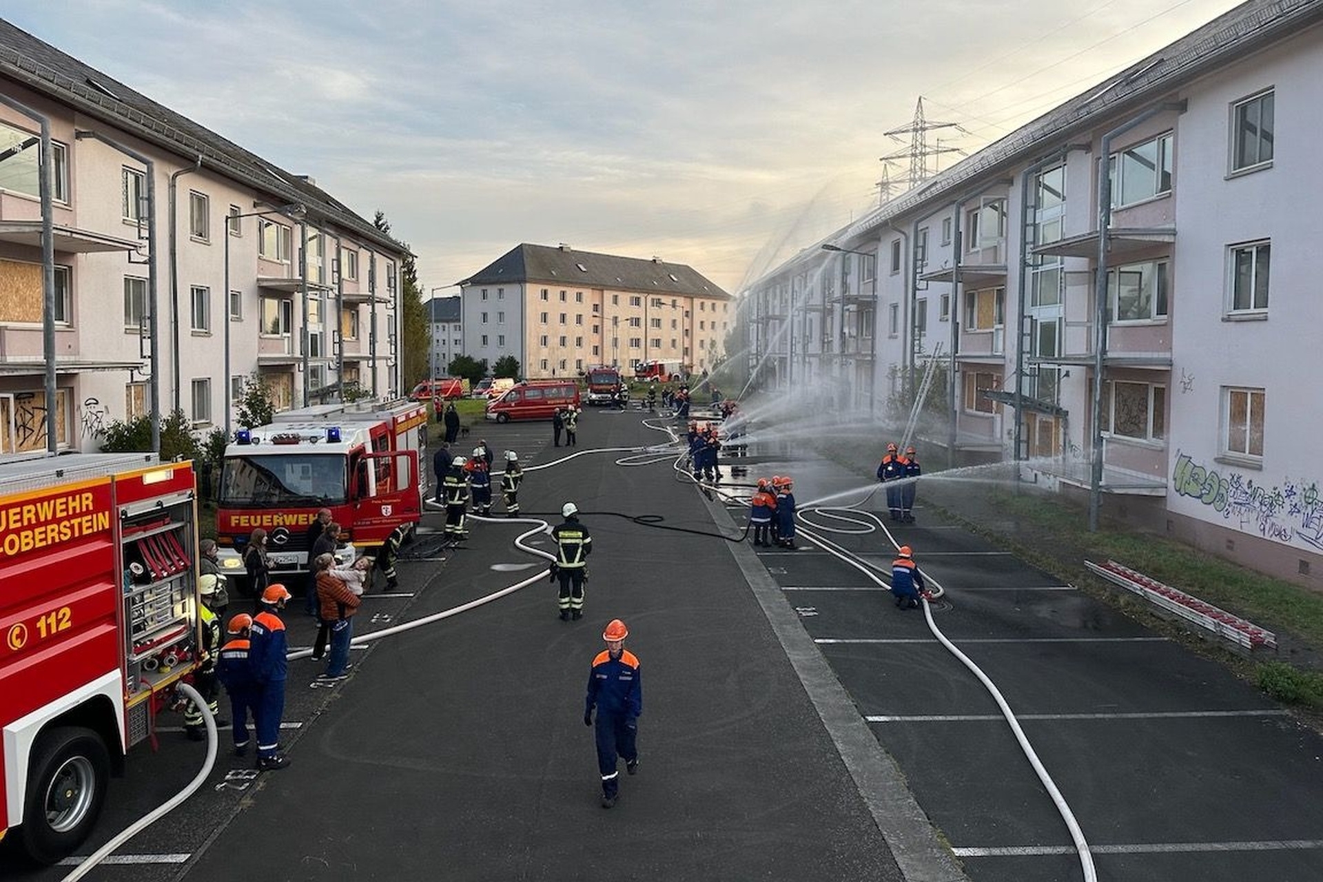 The photo shows a row of houses. In front of it are fire engines and members of the youth fire department, spraying water onto the houses with jet pipes.