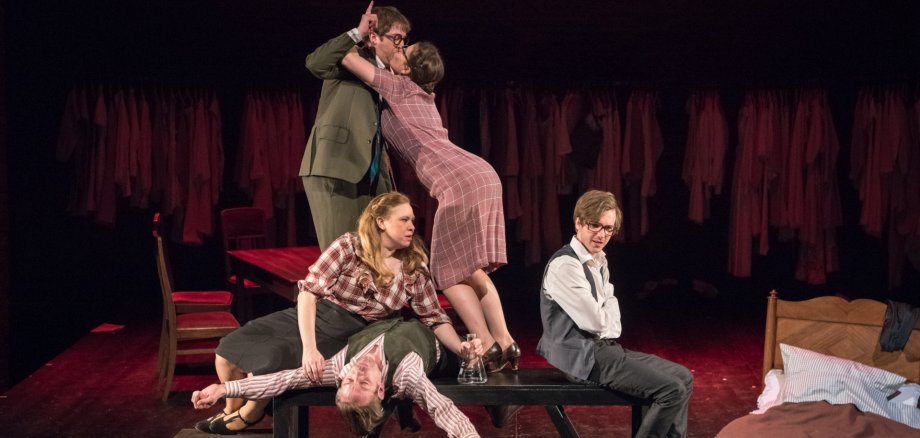 The photo shows five performers on a table. One is sitting with his arms folded and his back to the others. A man is lying on the table, apparently drunk, and a woman is leaning over him. Another man and a woman are standing on the table and kissing.