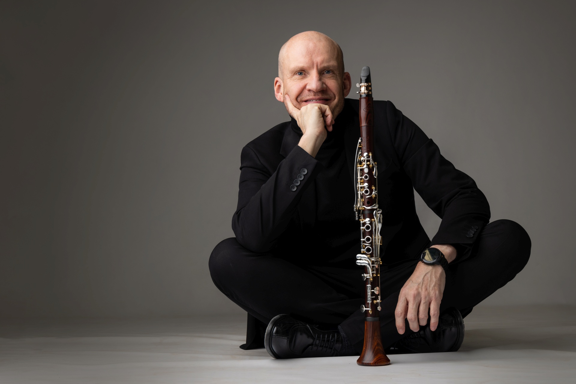 The photo shows the clarinettist sitting cross-legged on the floor in a black outfit. His instrument is on the floor in front of him.