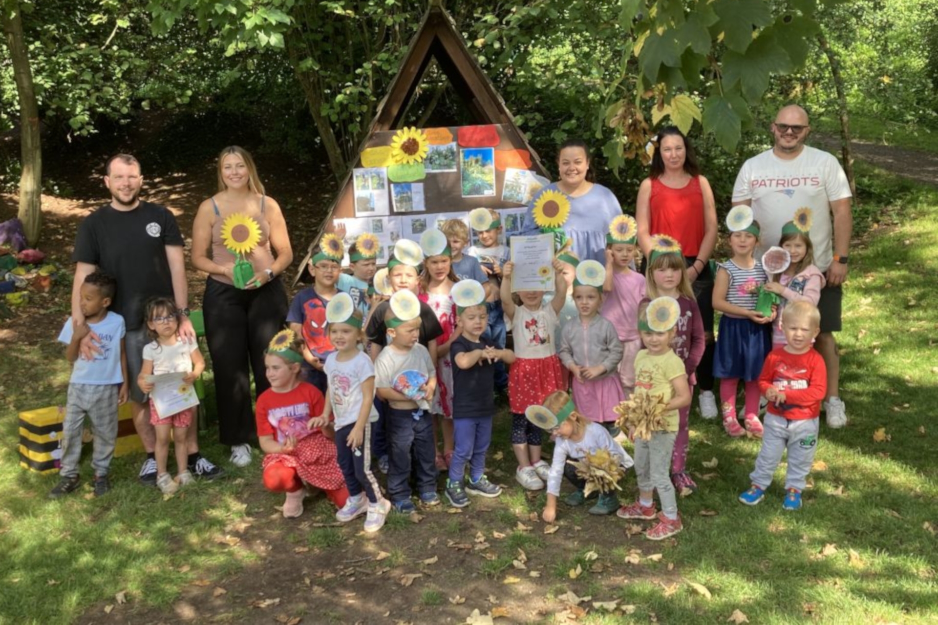 The photo shows children and adults. They are standing with sunflowers in a meadow in front of a wooden stand, with trees in the background.