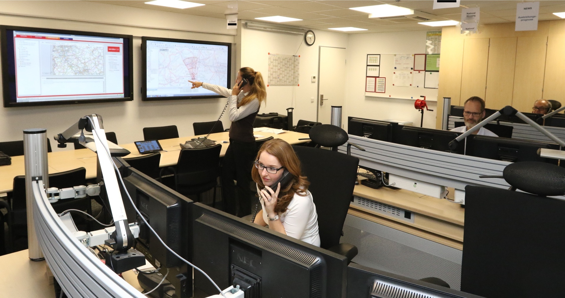 The photo shows a view into the BBK warning center, a woman sits in front of a screen and talks on the phone, a woman talks on the phone and points to a large screen on the wall, a man looks at his screens.