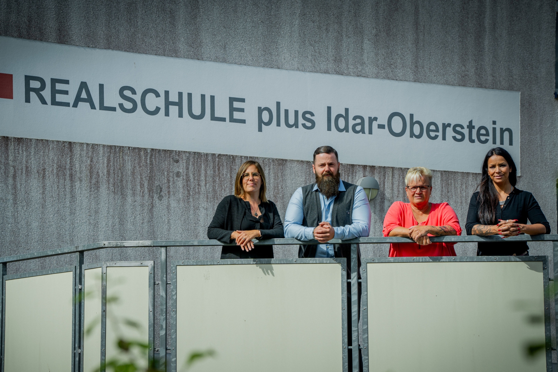 The photo shows the people mentioned leaning in a row in front of a sign with the inscription "Realschule plus Idar-Oberstein" on a railing.