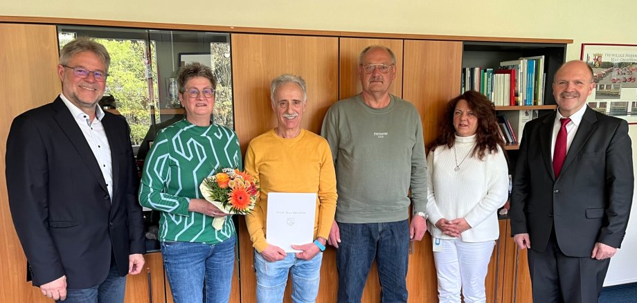 The photo shows six people, including Mr. Lange, who is holding the certificate of thanks, and his wife, who is holding a bouquet of flowers. The people are standing next to each other in front of a wall unit and looking into the camera.