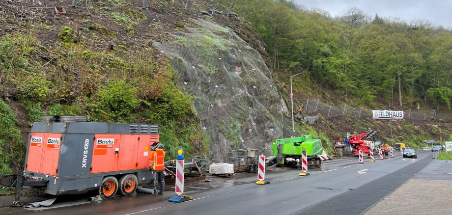 The photo shows a view of the rock on Mainzer Straße. Construction machinery can be seen in the foreground. Workers are installing a safety net above it.