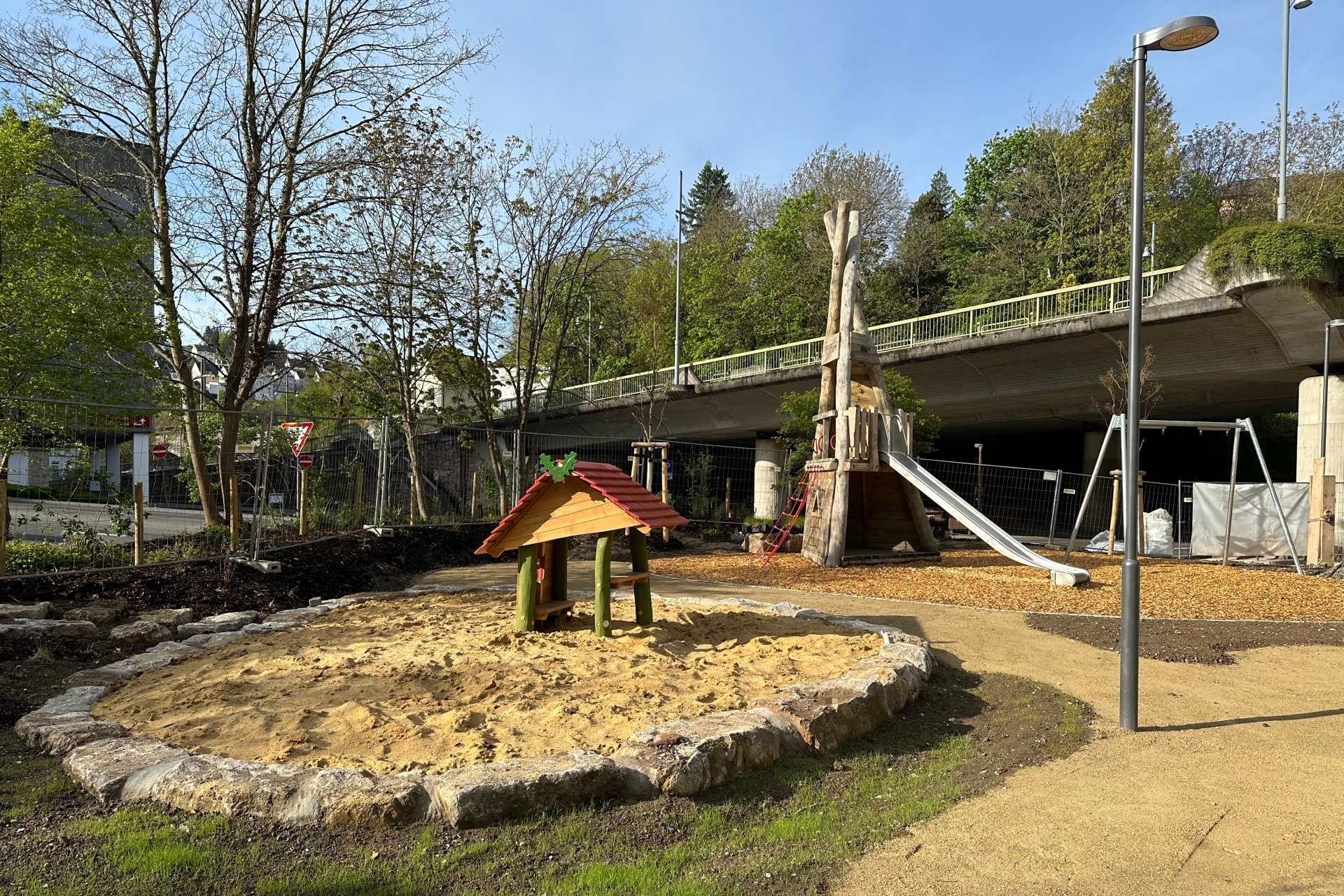 The photo shows a view of the playground. In the foreground, you can see a sandpit with a wooden house, in the background a wooden climbing frame with a slide and a swing.