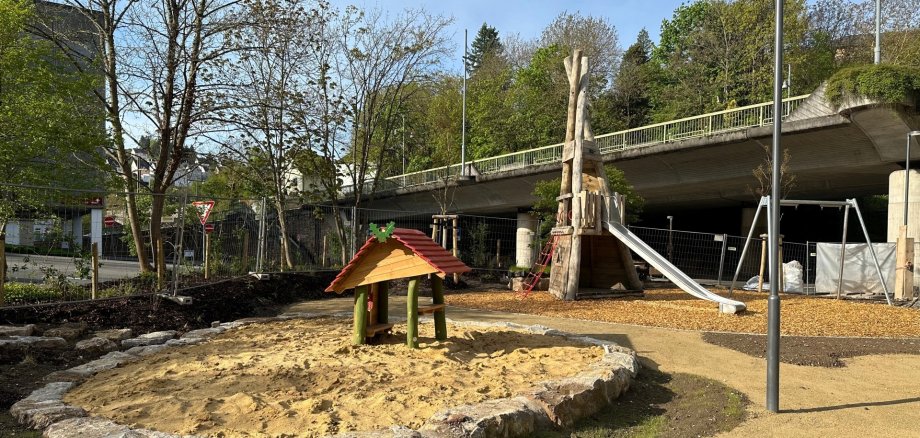 The photo shows a view of the playground. In the foreground, you can see a sandpit with a wooden house, in the background a wooden climbing frame with a slide and a swing.