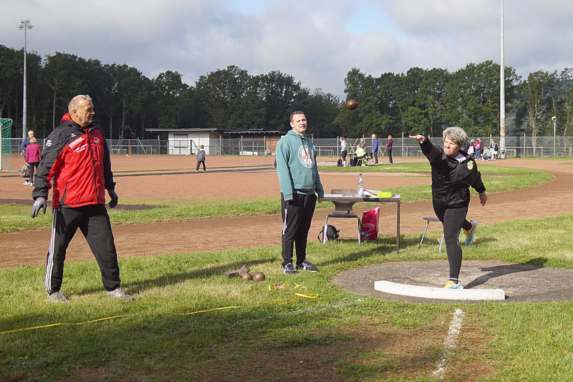 The photo shows a woman in a tracksuit throwing a shot put. Two judges are watching her.