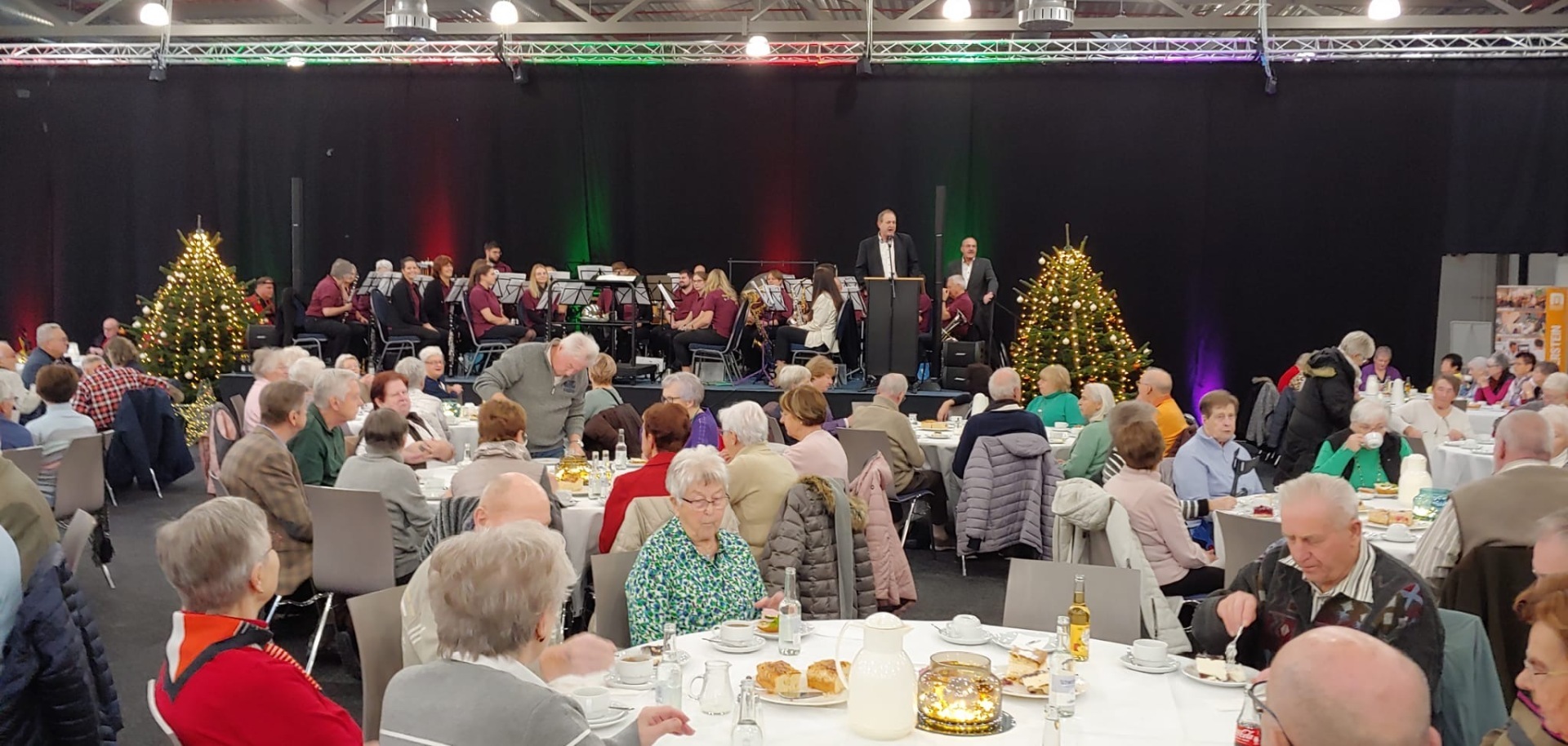 The photo shows a view of the exhibition hall with numerous guests at tables. Members of the Tiefenstein Music Society are sitting on the stage in the background. Mayor Frühauf is at the microphone, with Karl-Heinz Totz next to him.