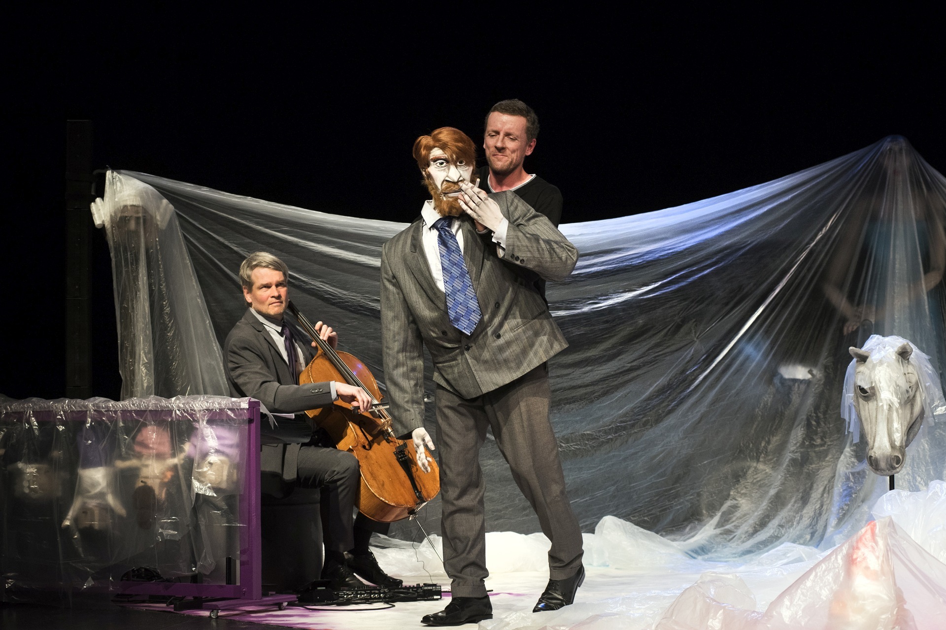 The photo shows an actor with a life-size figure on a stage. Behind him is a cellist sitting on a stool.
