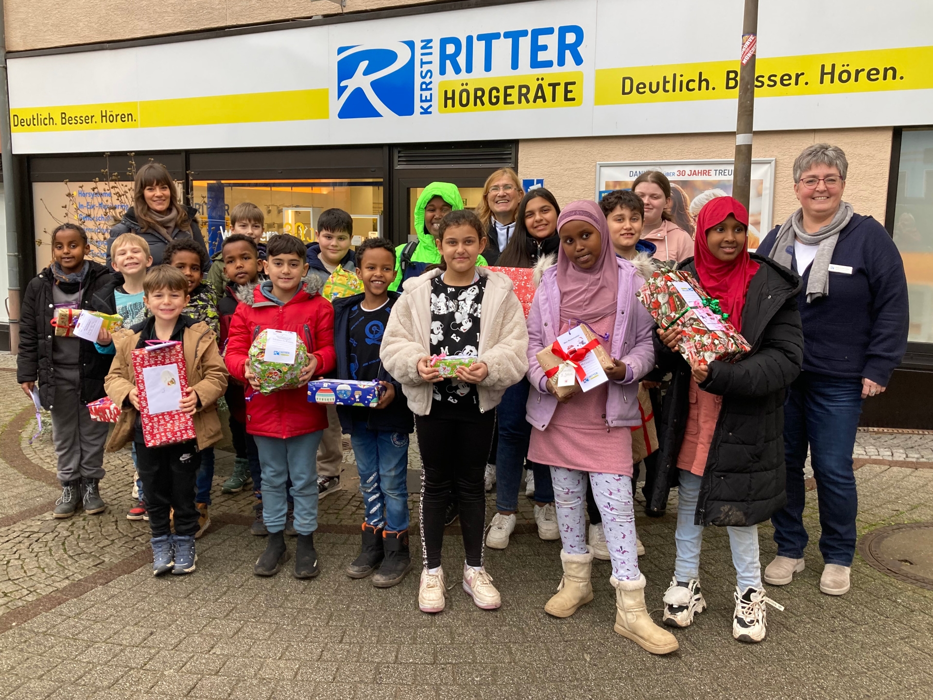 The photo shows the children and supervisors of the play and learning center as well as the store employees. They are standing in front of the store and the children are holding the presents in their hands.