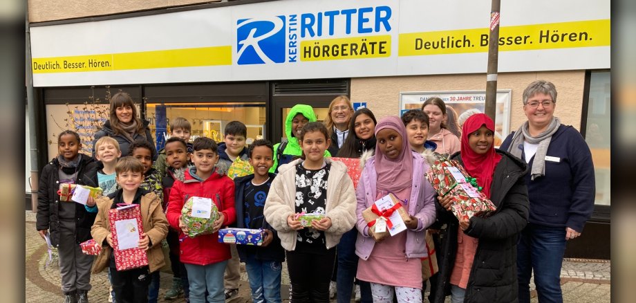 The photo shows the children and supervisors of the play and learning center as well as the store employees. They are standing in front of the store and the children are holding the presents in their hands.