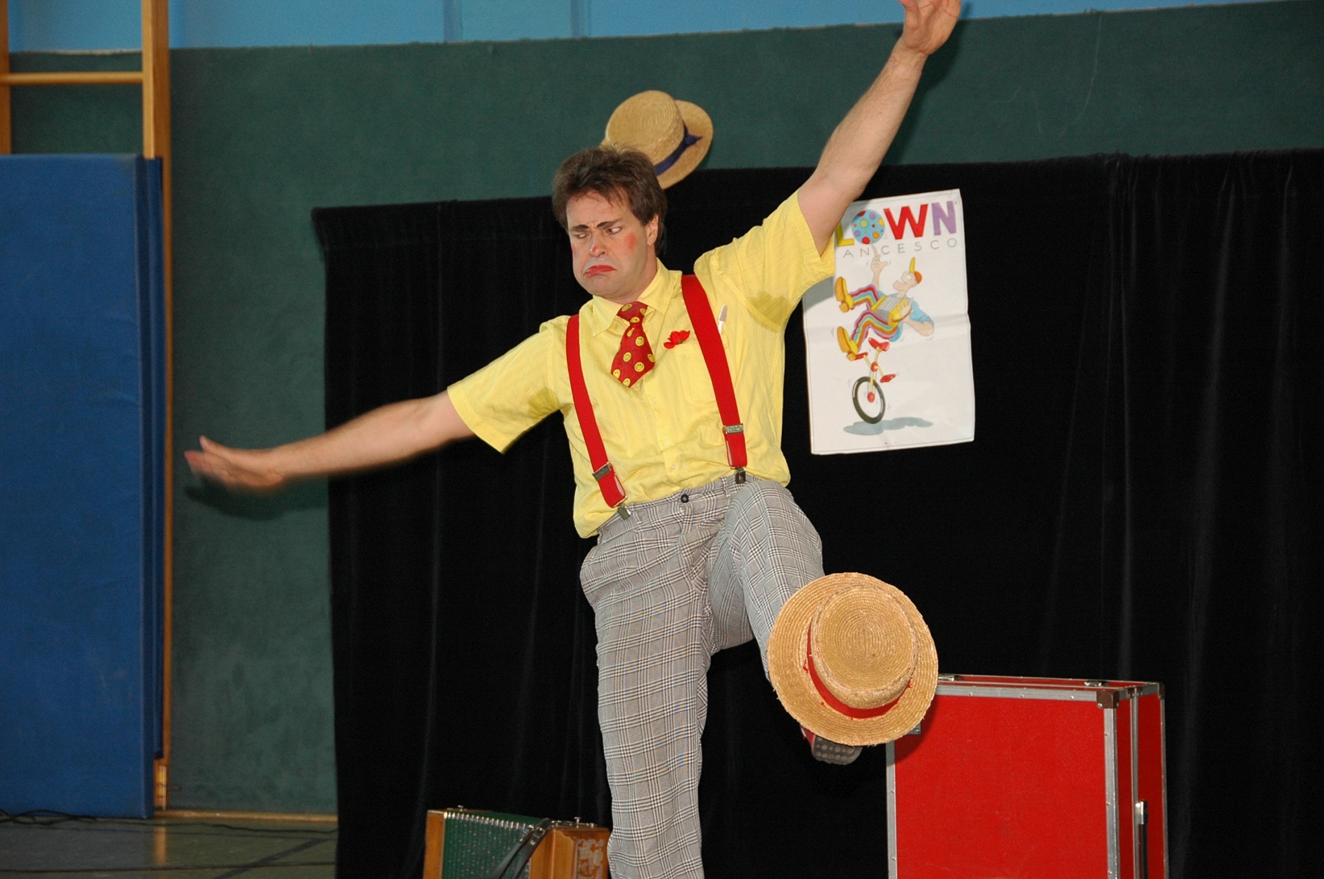 The photo shows a man in clown costume balancing on his right leg and juggling a hat at the foot of his left leg.
