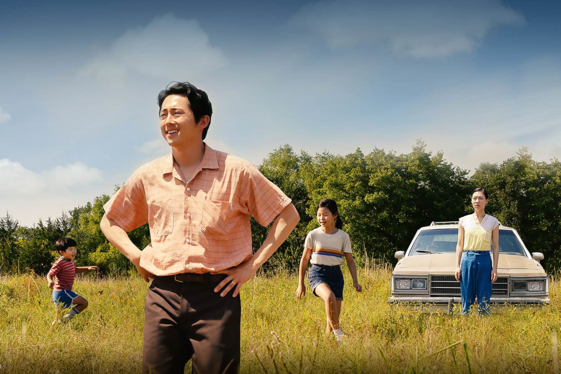 The photo shows a family of four, the parents with a daughter and a son. They are standing in a meadow with tall grass, behind them is their car. Father and mother are looking into the distance, the children are jumping around.