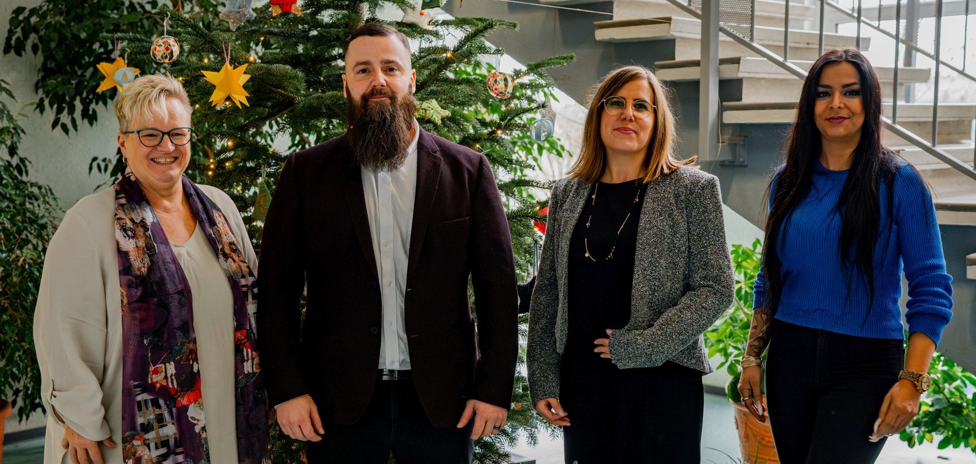 The photo shows the aforementioned people standing next to each other in front of a Christmas tree and looking into the camera.