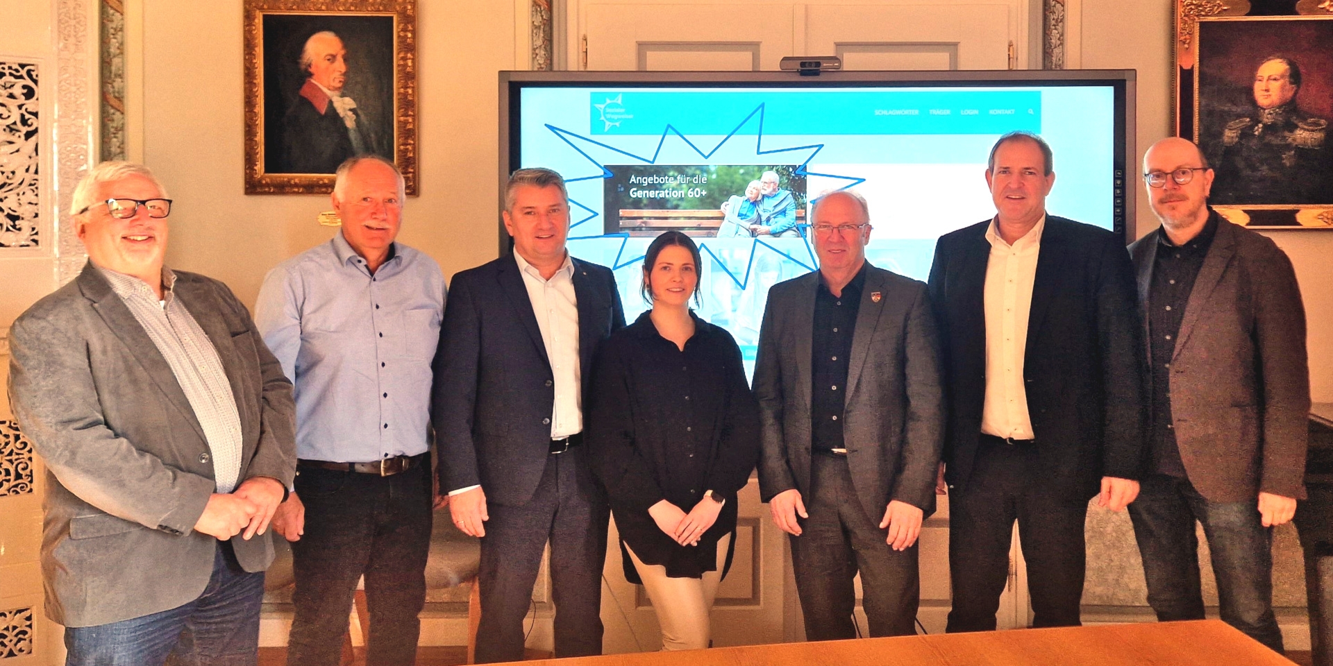 The photo shows the people mentioned standing next to each other in front of a screen and looking into the camera. The screen shows the website with the senior citizens' platform.