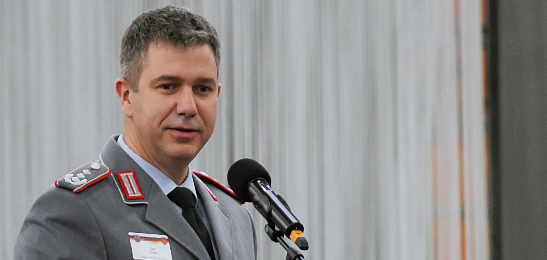 The photo shows a close-up of Colonel Tuneke standing on the stage at the lectern.