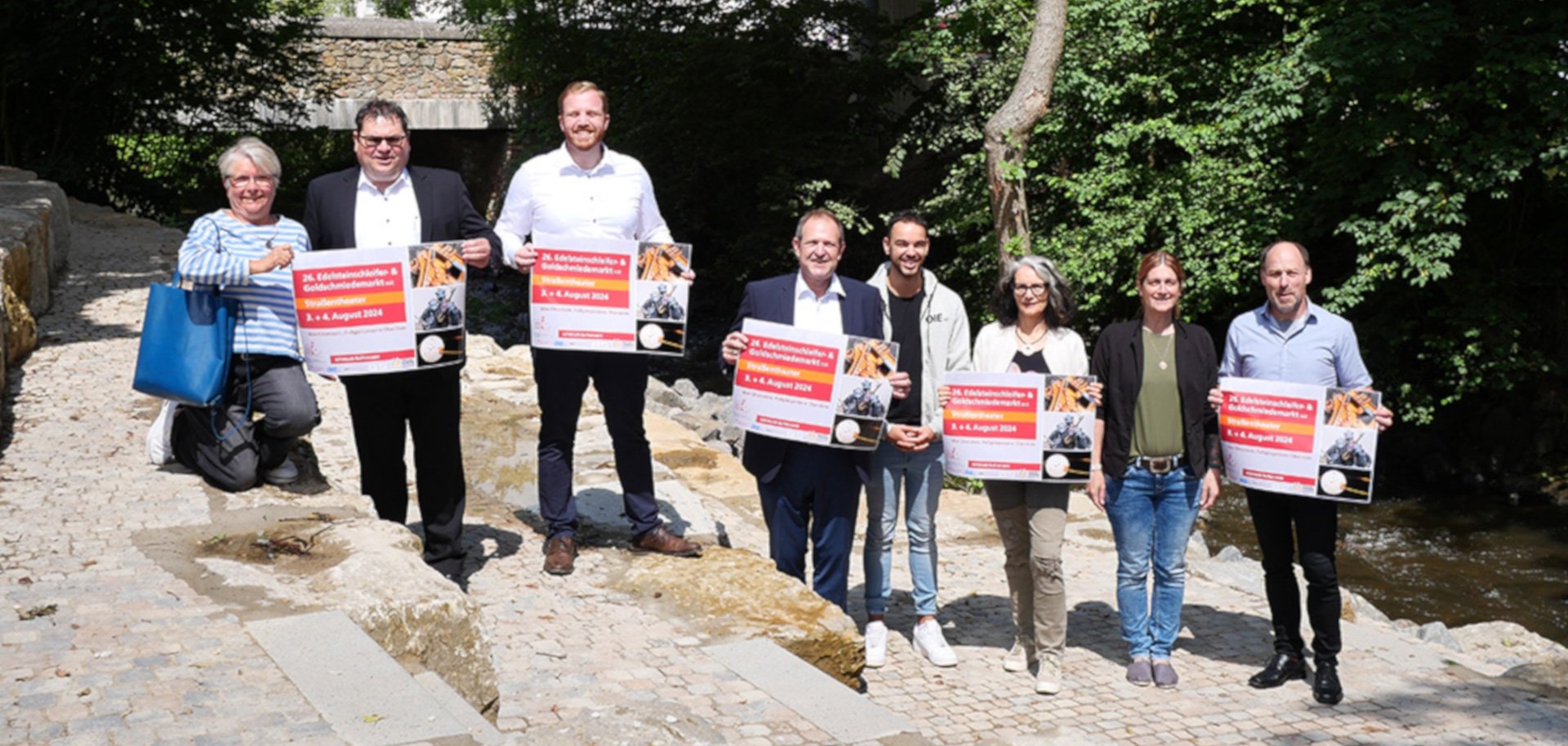 The photo shows the organizers and sponsors standing on the stone steps of the Helmut-Kohl-Europaplatz on the banks of the Idarbach. They are holding event posters in their hands and looking into the camera.