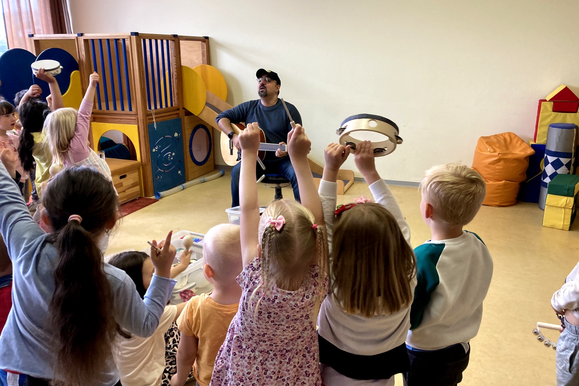 The photo shows the view into the movement room during the "Music Mice" activity. In the background, Mike Stauder sits on a chair with a guitar and sings. The children are dancing in the foreground.