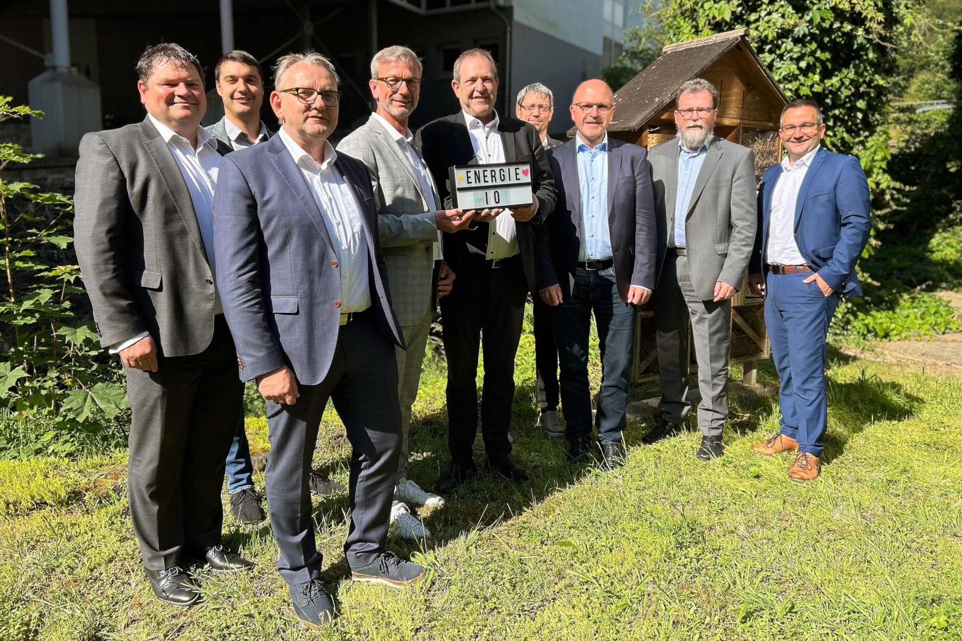 The photo shows the people mentioned in the text. They are standing next to each other on a meadow and looking into the camera. Mr. Johann and Mayor Frühauf are holding a sign with the inscription Energie IO.