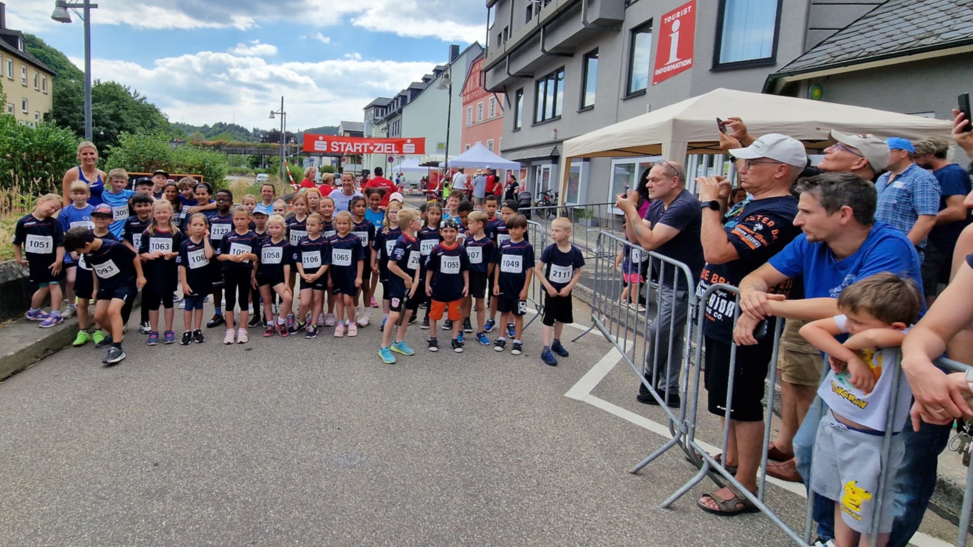The photo shows many children with start numbers standing in the start-finish area waiting for the starting signal. To the side you can see parents with cell phones taking photos.