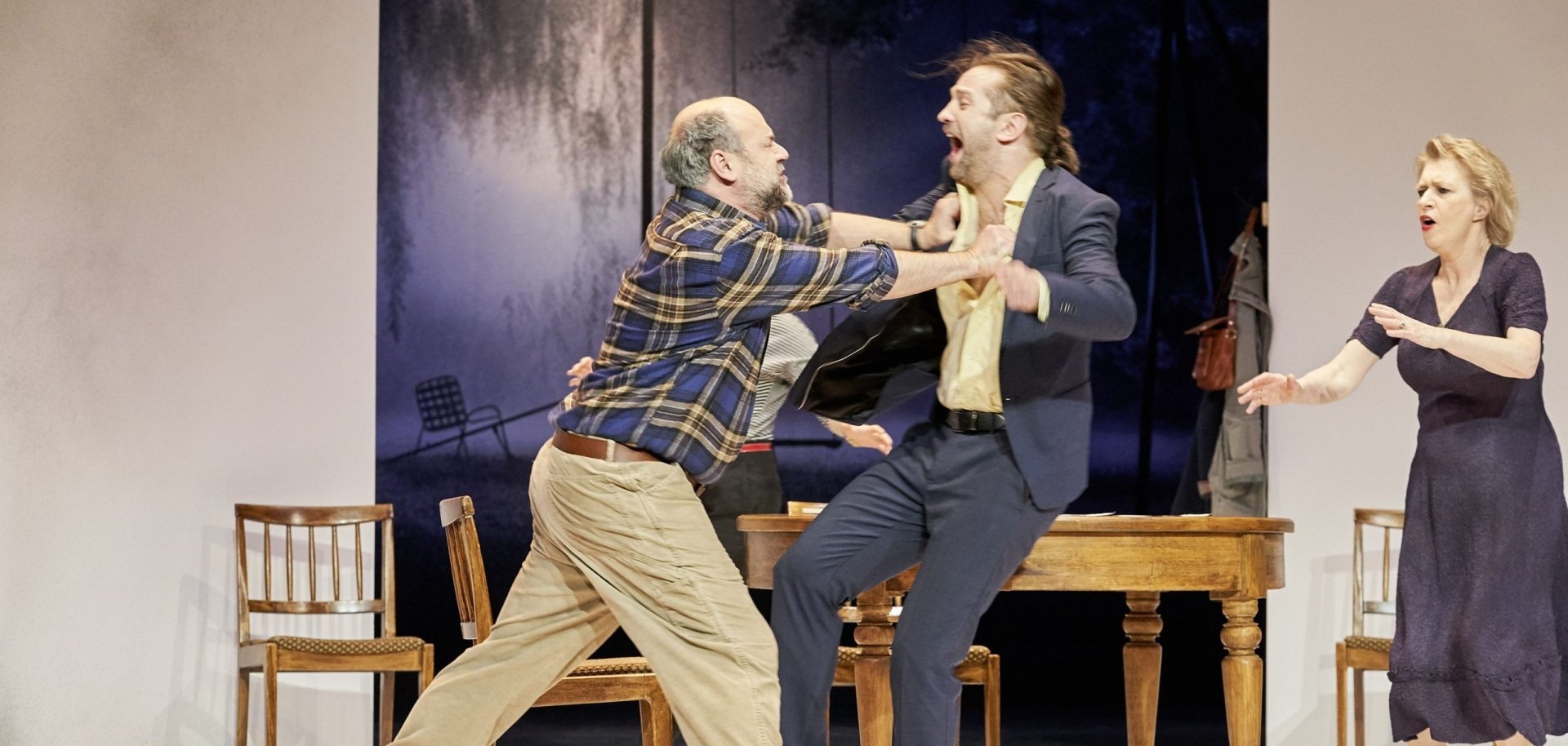 The photo shows three actors, two men and a woman. The two men are fighting with each other, the woman is looking at them in horror.