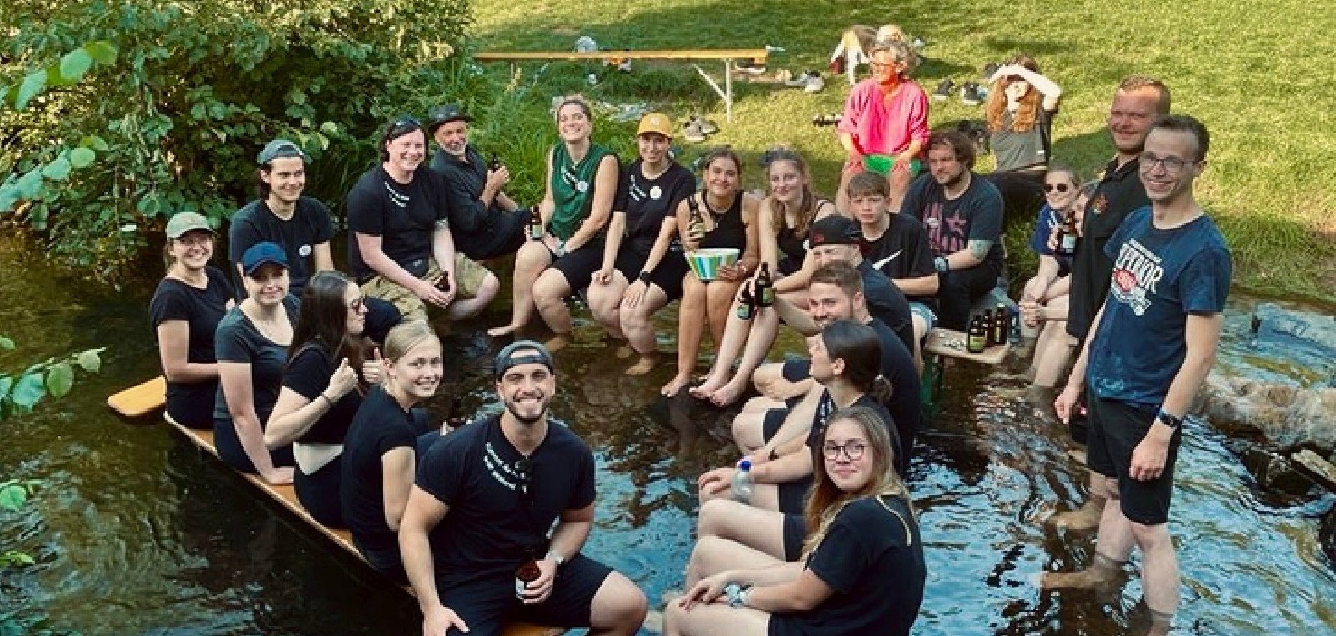 The photo shows a large group of helpers. They are sitting on beer benches in the Idarbach and laughing into the camera.