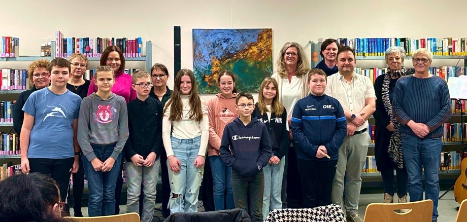 The photo shows participants, organizers and jury of the district competition. They stand next to each other in front of a wall in the city library and look into the camera,
