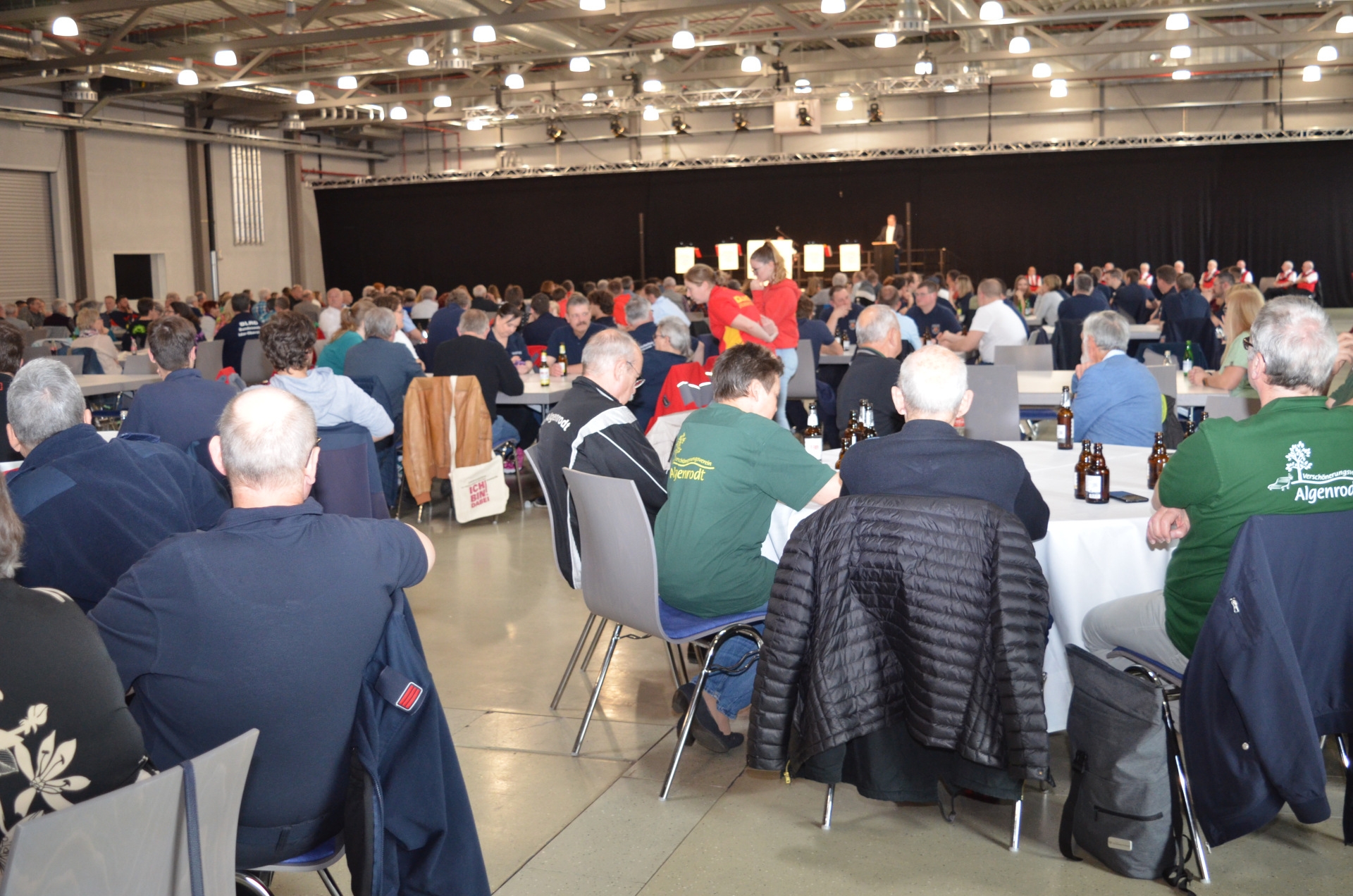 The photo shows a view of the exhibition hall. You can see many people sitting at round tables, with drinks on the tables. In the background you can see the stage on which Lord Mayor Frank Frühauf is giving a speech.