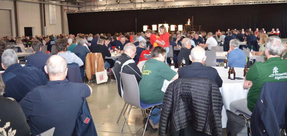 The photo shows a view of the exhibition hall. You can see many people sitting at round tables, with drinks on the tables. In the background you can see the stage on which Lord Mayor Frank Frühauf is giving a speech.