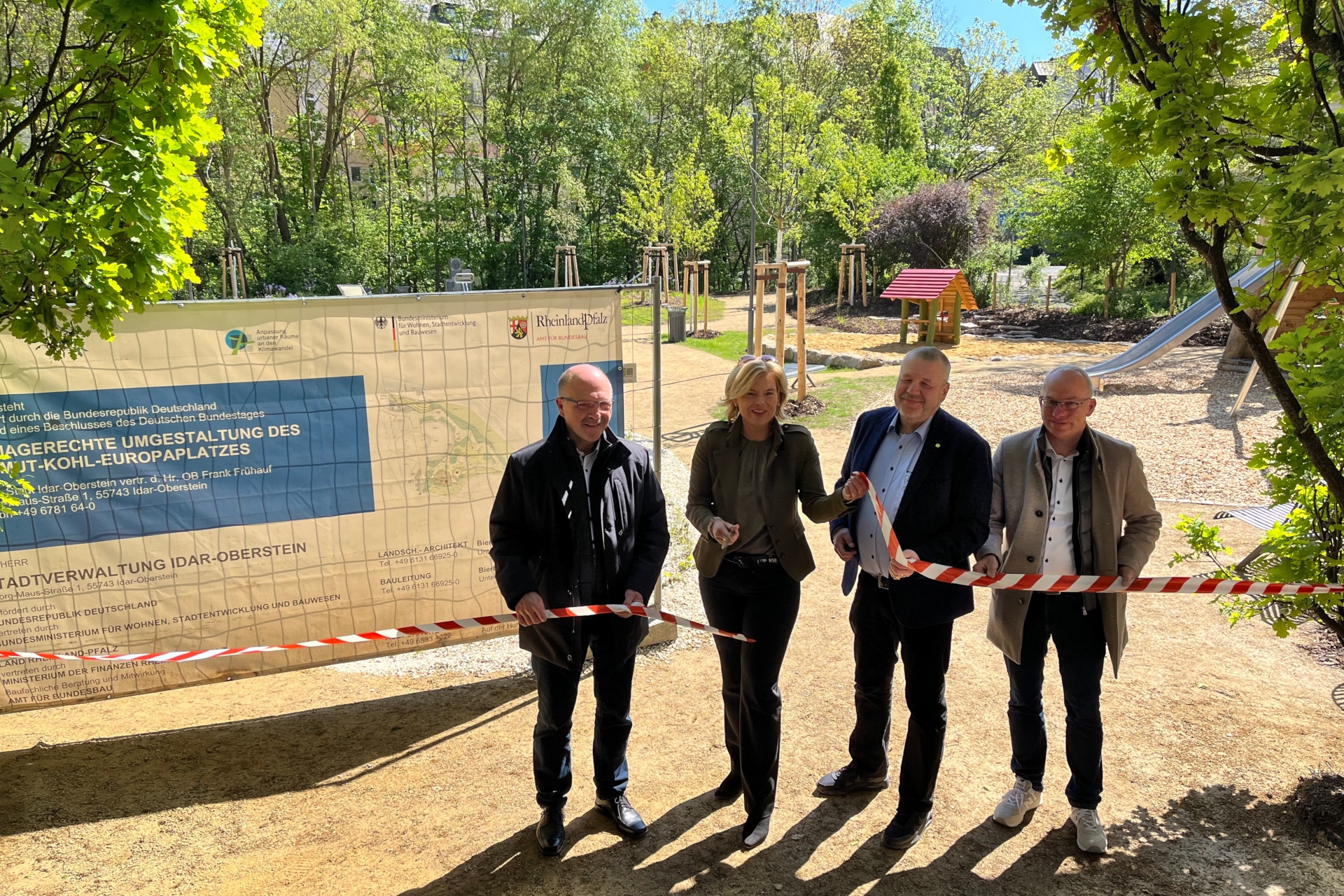 The photo shows the people mentioned. They are standing next to each other behind a track tape and looking into the camera, Julia Klöckner has a pair of scissors in her hand and has just cut the tape. In the background you can see the playground area.