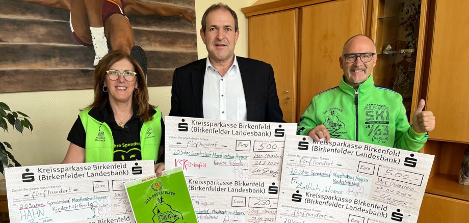 The photo shows the people mentioned. They are standing next to each other in the mayor's office, holding donation cheques in their hands and looking into the camera.